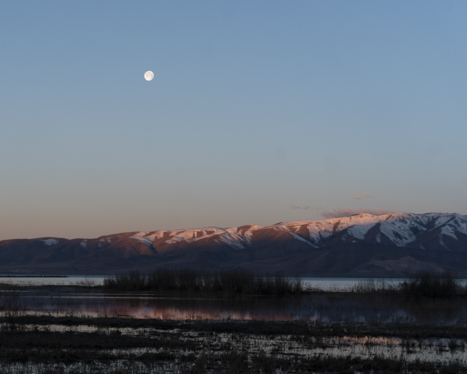 Snowy mountains across the lake catch the first days sun, the moon above